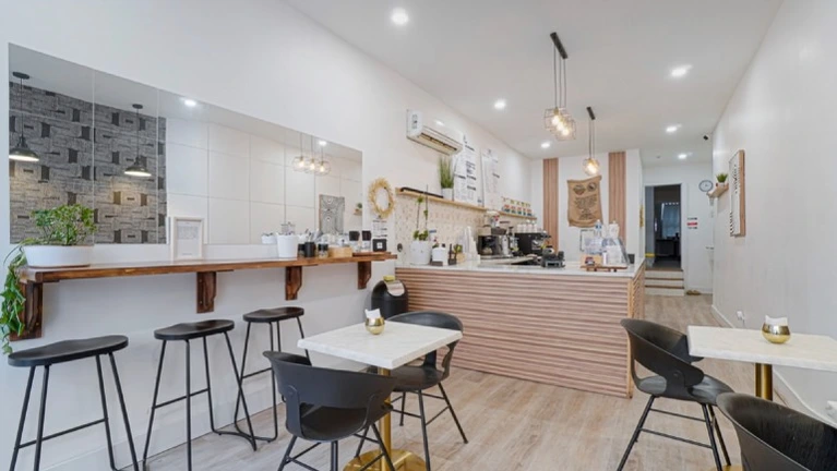 Bright customer seating area, featuring small marble-top tables with black modern chairs, a wooden bar counter with stools along a mirrored wall