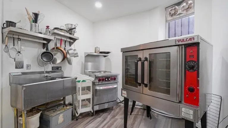 Compact commercial kitchen area, equipped with a large stainless steel sink, a pot rack, a small range, and double-stacked Vulcan convectio